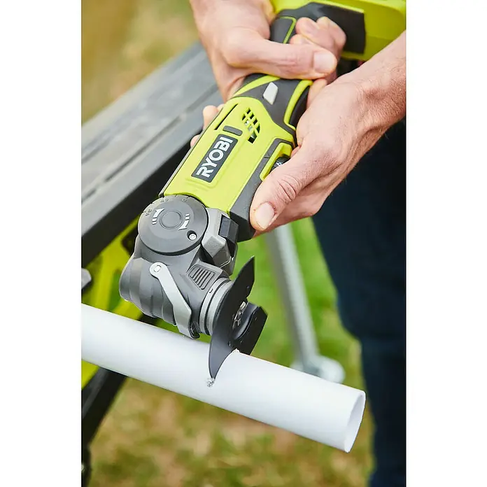 Ryobi ONE+ Maschinenpaket - Akku-Multifunktionswerkzeug R18MT-0 + gratis Starter-Kit - Nahaufnahme einer Ryobi Oszillationssäge, die ein weißes PVC-Rohr durchschneidet, gehalten von Händen mit blauen Hosen im Hintergrund.