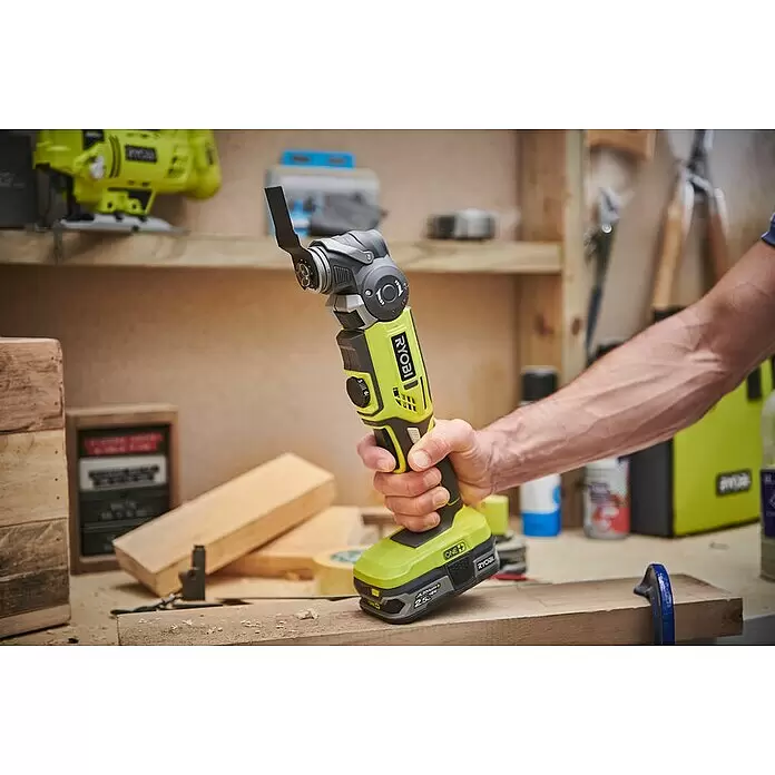 Ryobi ONE+ Maschinenpaket - Akku-Multifunktionswerkzeug R18MT-0 + gratis Starter-Kit - Eine Hand hält ein limettengrünes und schwarzes Ryobi Multifunktionswerkzeug mit Klinge über einer Holzoberfläche in einer Werkstatt.