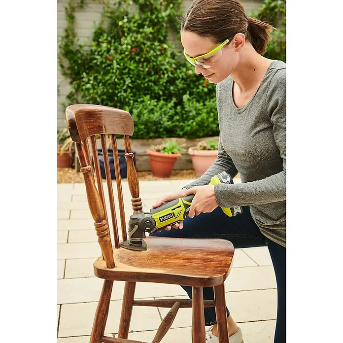 Ryobi ONE+ Akku-Multifunktionswerkzeug R18MT-0 - 18 V, Ohne Akku - Eine Frau schleift einen Holzstuhl mit einem gelben Ryobi-Multifunktionswerkzeug im Freien, Schutzbrille trägt.
