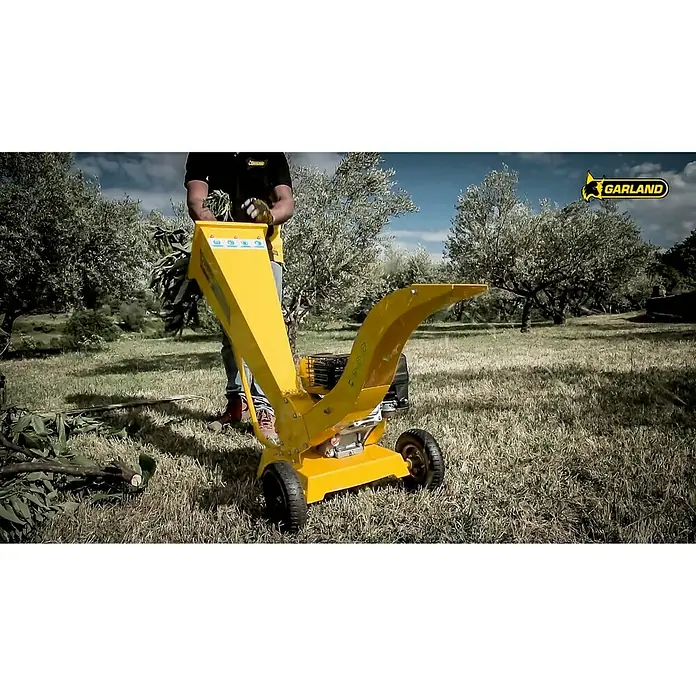 Garland Biotrituradora de gasolina 780 QG-V17 - Grosor de rama máx.: 6 cm, 4,3 kW, Cilindrada: 212 cm³ - Una astilladora amarilla Garland en acción, triturando ramas y ramitas, con operador y fondo de olivos.