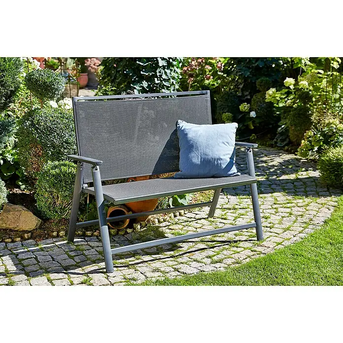 Sunfun Maja Gartenbank - B x T x H: 119 x 67 x 96 cm, Textilen, Anthrazit, Klappbar - Eine stilvolle graue Gartenbank mit einer blauen Kissenauflage steht auf einem gepflasterten Weg in einem üppigen Garten.