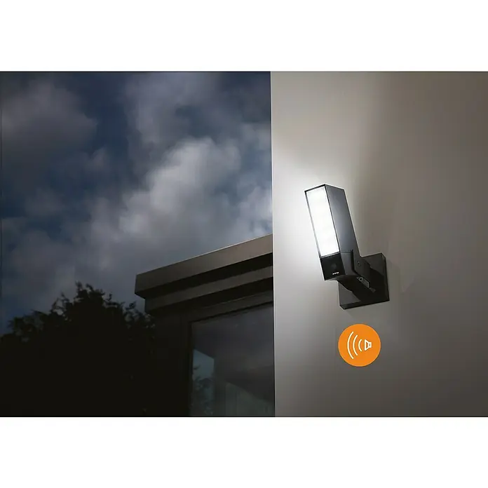 Netatmo Cámara de vigilancia con sirena - Negro, Ángulo de detección: 100 °, Alcance de detección: 15 m - Imagen de una moderna lámpara de pared exterior negra con luz brillante, montada en una pared junto a un edificio al atardecer.