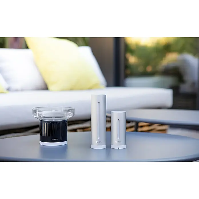 Netatmo Estación meteorológica con pluviómetro inteligente - Pack módulo interior + pluviómetro inteligente - Primer plano de tres dispositivos de estación meteorológica Netatmo en una mesa al aire libre.