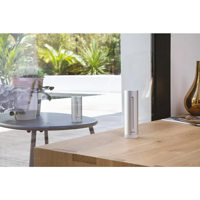 Netatmo Estación meteorológica con pluviómetro inteligente - Pack módulo interior + pluviómetro inteligente - Primer plano de un módulo interior de la estación meteorológica Netatmo sobre una mesa de madera, con otro módulo y plantas de interior al fondo.