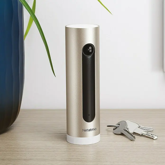 Netatmo Cámara de vigilancia Welcome - Marrón, Ángulo de detección: 130 ° - Primer plano de una cámara de seguridad Netatmo en oro y blanco sobre una mesa con llaves y una planta.