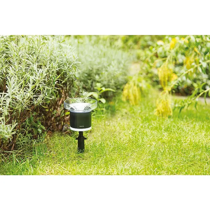 Netatmo Soporte - Específico para: Pluviómetro o pluviómetro Netatmo, L x An x Al: 12,7 x 7,62 x 10,16 cm - Primer plano de un pluviómetro Netatmo en un jardín, rodeado de plantas verdes. Medición precisa para tu jardín con este dispositivo inteligente.