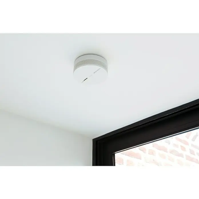 Netatmo Detector de humo inteligente - Diámetro: 11,5 cm, Duración de batería: 10 años, Señal de alarma: 85 dB - Un detector de humo redondo y blanco instalado en un techo blanco en una habitación luminosa, ofreciendo seguridad.