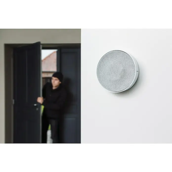 Netatmo Sirena interior - Específico para: Cámara Interior Inteligente Netatmo - Alarma antirrobo de pared con un ladrón entrando por la puerta al fondo. El enfoque está en la alarma redonda y blanca con una malla fina.