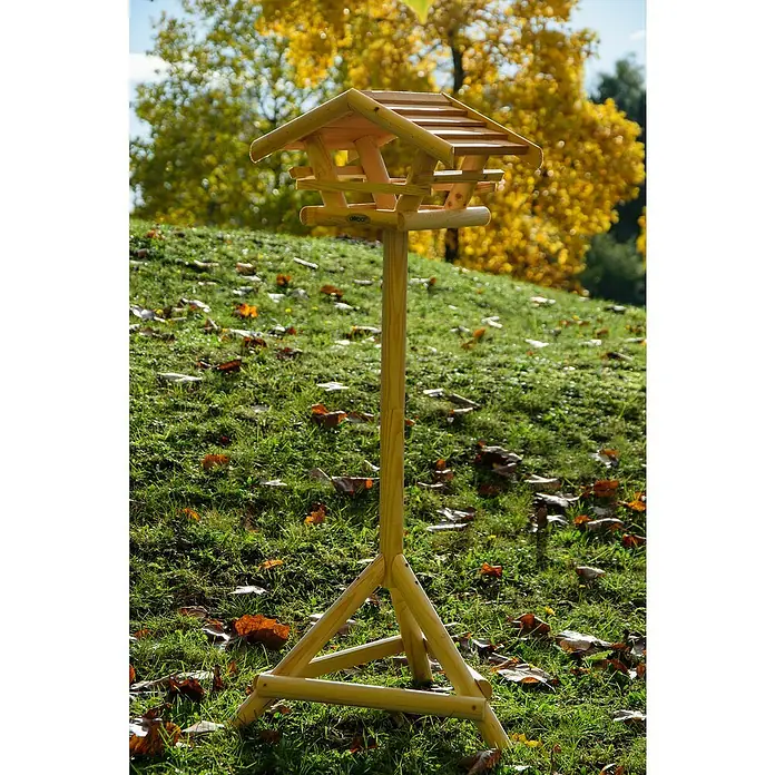 Dobar Vogelhaus Bernie Bernstein - 44 x 44 x 101 cm, Holz, Braun - Ein hölzernes Vogelhaus auf einem grünen Hügel mit gelben Blättern im Hintergrund. Das Vogelhaus hat ein Dach und steht auf einem dreibeinigen Ständer.
