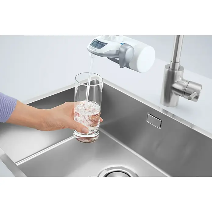 Brita Filtro de agua On Tap - Caudal: 1,6 l/min - Un filtro de agua Brita llena un vaso con agua filtrada sobre un fregadero de acero inoxidable para garantizar agua potable.