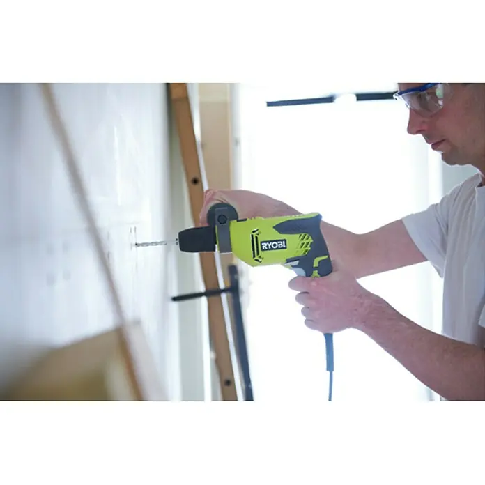 Ryobi Taladro percutor RPD500-G - 500 W, Número de revoluciones de marcha en vacío 1ª marcha: 3.000 r.p.m., Número máx. de impactos: 50.000 /min - Un hombre hace un agujero en la pared con un taladro Ryobi amarillo. La atención se centra en el taladro en acción.