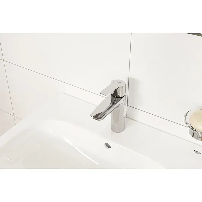 Grohe Start Grifo de lavabo M - Cromo, Resplandeciente, Barra de tracción - Primer plano de un grifo Grohe sobre un lavabo blanco con fondo de azulejos blancos.