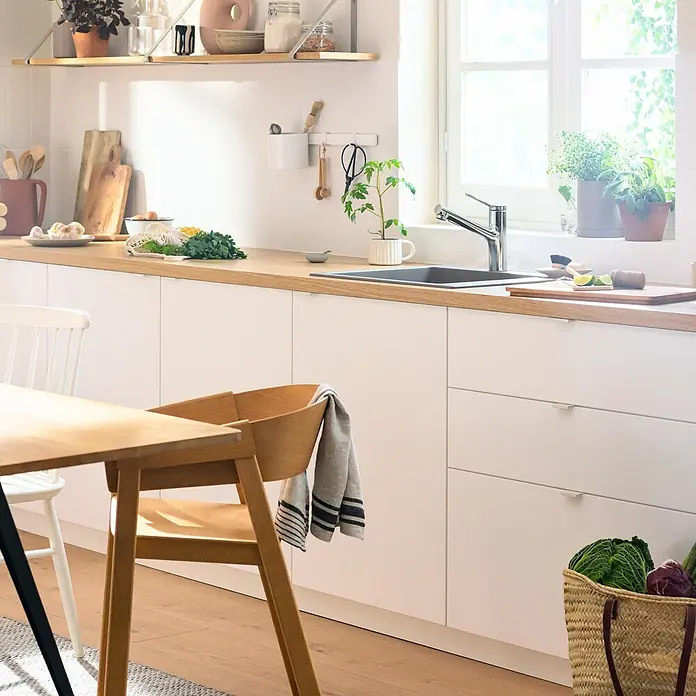 Hansgrohe Grifo de cocina Zesis - Cromo, Brillante, Extensible - Cocina luminosa con fregadero y grifo, encimera de madera, armarios blancos y estanterías. Hierbas frescas y utensilios de cocina a la vista.