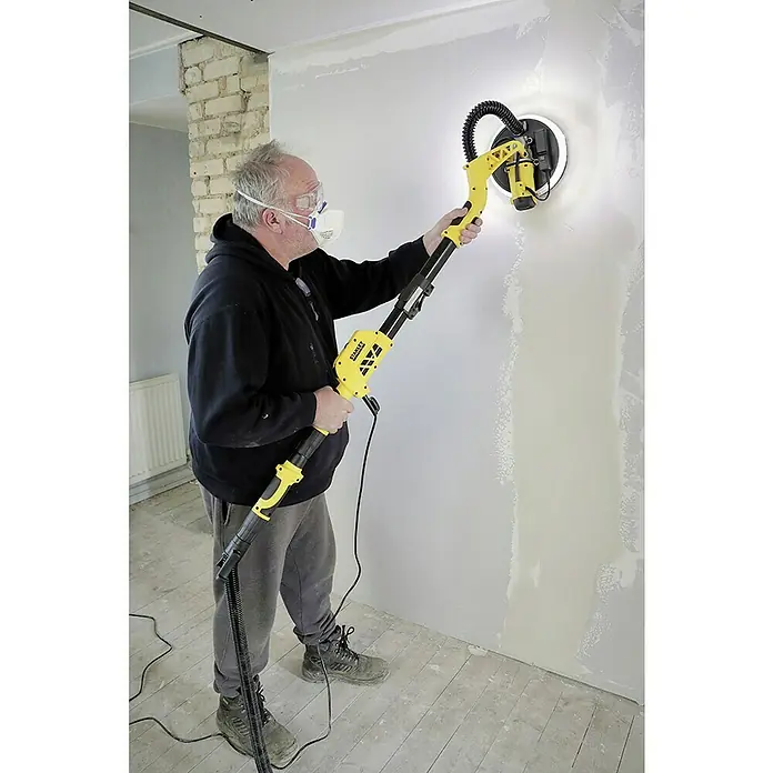 Stanley FatMax Lijadora de pared y techo SFMEE500S-QS - 750 W, Diámetro disco de lija: 225 mm - Un hombre usa una lijadora de pared amarilla Stanley para alisar una pared mientras usa gafas de seguridad y una máscara.
