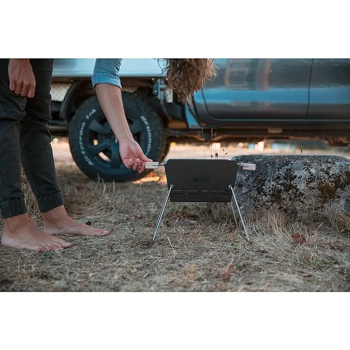 Knister Holzkohlegrill - Grillfläche (B x T): 28 x 20 cm, Schwarz - Eine Person, die einen tragbaren Krister Grill im Freien benutzt. Der Grill steht auf trockenem Gras neben einem Felsen und einem Auto.
