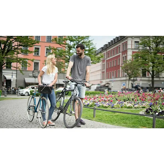 Knister Fahrradhalterung - L x B x H: 13 x 13 x 10 cm, Passend für: Knister Grill Modelle / Handelsübliche Fahrradlenker - Ein Paar steht mit Fahrrädern auf einem gepflasterten Weg, im Hintergrund Gebäude und Blumenbeete.