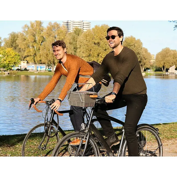 Knister Fahrradhalterung - L x B x H: 13 x 13 x 10 cm, Passend für: Knister Grill Modelle / Handelsübliche Fahrradlenker - Zwei Männer radeln an einem sonnigen Tag am See entlang, einer hat einen auffälligen Fahrradkorb, der die Aufmerksamkeit auf sich zieht.