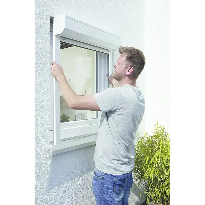 Schellenberg Rollladen-Komplett-Set MINI - Vorbau, 80 x 100 cm - Ein Mann montiert ein Rollladensystem an einem Fenster, wobei die weiße Farbe des Rollladens und des Rahmens hervorsticht.