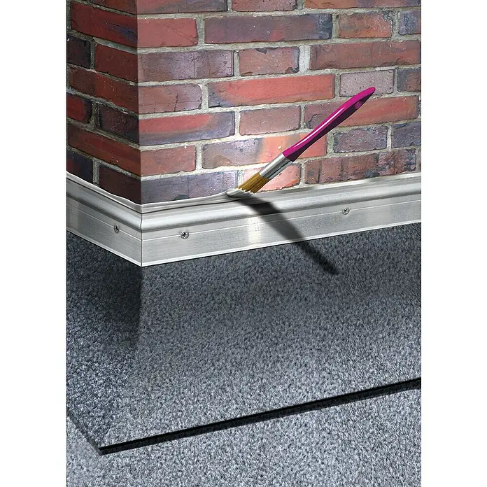 MEM Dicht-Fix - 4 l - Farbpinsel mit pinkfarbenem Griff, der weiße Farbe auf eine Aluminiumfußleiste aufträgt, vor einem Hintergrund aus Ziegelsteinen und Teppich.