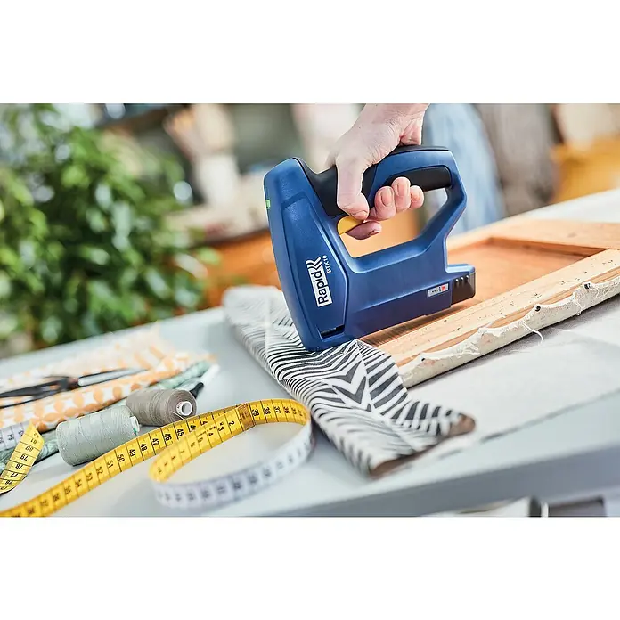 Rapid Akku-Tacker BTX10 - 3,6 V, Klammertiefe: 4 mm - 10 mm - Nahaufnahme eines Rapid BTX10 Akku-Tackers beim Befestigen von Stoff an einem Holzrahmen, umgeben von Nähzubehör auf einem Tisch.
