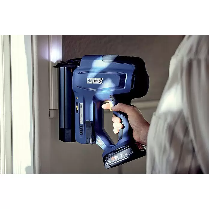 Rapid Power for All 18V Akku-Nagler BNX50 - 18 V, Ohne Akku, Länge Nagel: 20 mm - 50 mm - Eine Hand hält eine blaue Rapid Akku-Nagelpistole mit 18V 2,5Ah Akku und drückt sie gegen eine weiße Oberfläche.