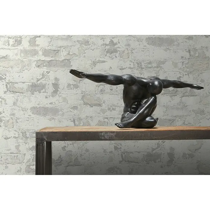 A.S. Création Metropolitan Stories Vliestapete Mauerwerk - Grau/Weiß, Steinoptik, 10,05 x 0,53 m - Eine bronzefarbene Skulptur eines sitzenden Mannes auf einem rustikalen Holztisch vor einer Ziegelwand.