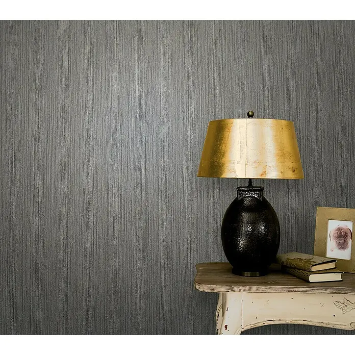 Marburg Modernista Vliestapete - Mittelgrau, Uni, 10,05 x 0,53 m - Dekorative Tischleuchte mit goldenem Schirm und dunklem Sockel, elegant auf einem Beistelltisch mit Büchern und Bilderrahmen platziert.