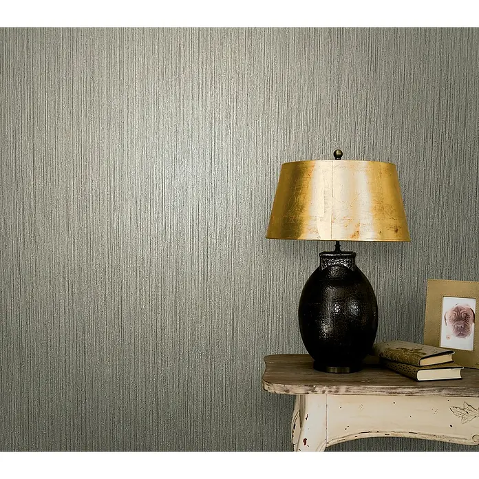 Marburg Modernista Vliestapete - Schlamm, Uni, 10,05 x 0,53 m - Elegante Tischleuchte mit goldenem Schirm auf einem Beistelltisch, vor einer strukturierten Wand für eine gemütliche Atmosphäre.