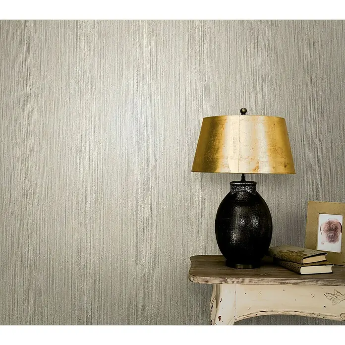 Marburg Modernista Vliestapete - Beige, Uni, 10,05 x 0,53 m - Elegante Innenaufnahme mit einer Tischlampe mit goldenem Schirm auf einem Beistelltisch, vor einer strukturierten Wand.