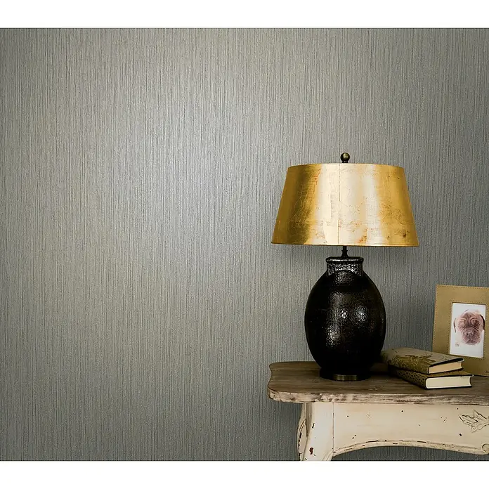 Marburg Modernista Vliestapete - Grau, Uni, 10,05 x 0,53 m - Elegante Tischleuchte mit goldenem Schirm und schwarzem Sockel, platziert auf einem Beistelltisch mit dekorativen Büchern und Fotorahmen.