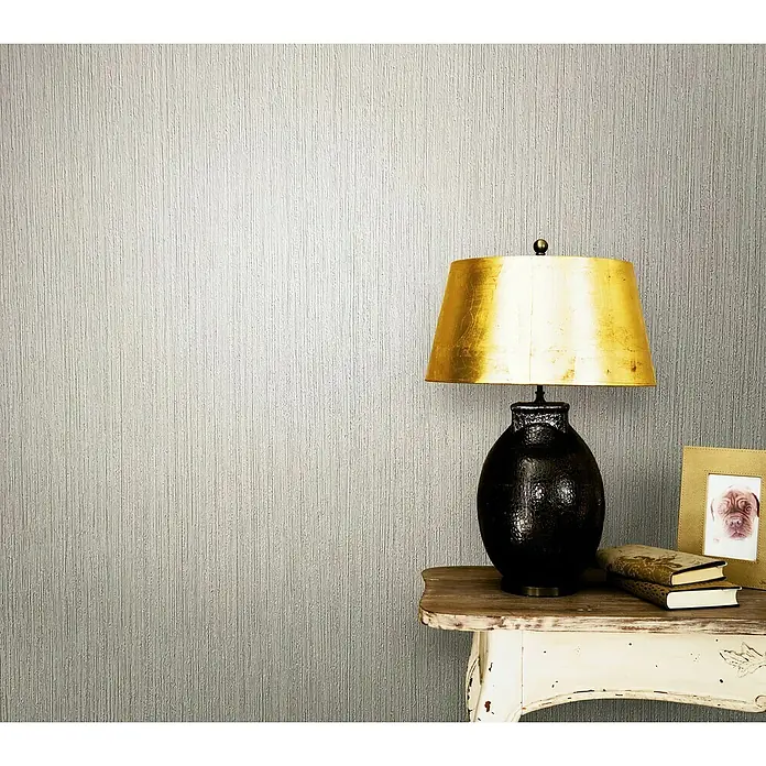 Marburg Modernista Vliestapete - Creme-Beige, Uni, 10,05 x 0,53 m - Elegante Tischlampe mit goldenem Schirm auf einem Beistelltisch, vor einer strukturierten Wand. Schafft eine warme und stilvolle Atmosphäre.