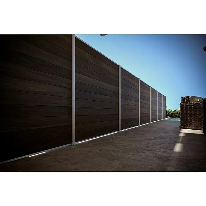 T & J Sichtschutzelement Elskop - B x H: 179 x 15 cm, Teak - Eine lange, dunkle Holzwand mit hellen Metallstützen trennt einen Außenbereich ab und bietet Privatsphäre. Der Boden ist aus dunklem Beton.