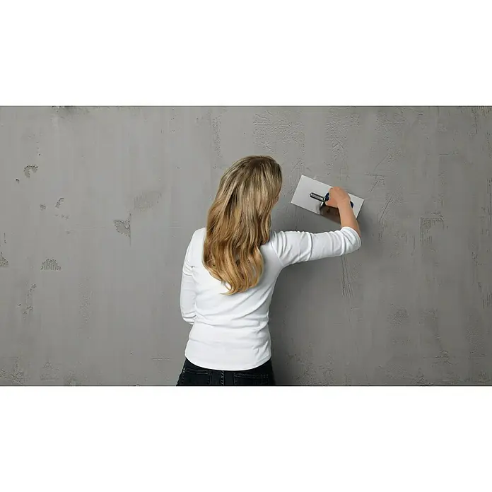 SCHÖNER WOHNEN-Farbe Trendstruktur Dekoputz-Set Sichtbeton-Optik - 3 -tlg. - Eine Frau glättet eine graue Wand mit einer Glättkelle, um eine gleichmäßige Oberfläche zu erzielen. Die Renovierungsarbeit verleiht dem Raum ein sauberes Aussehen.
