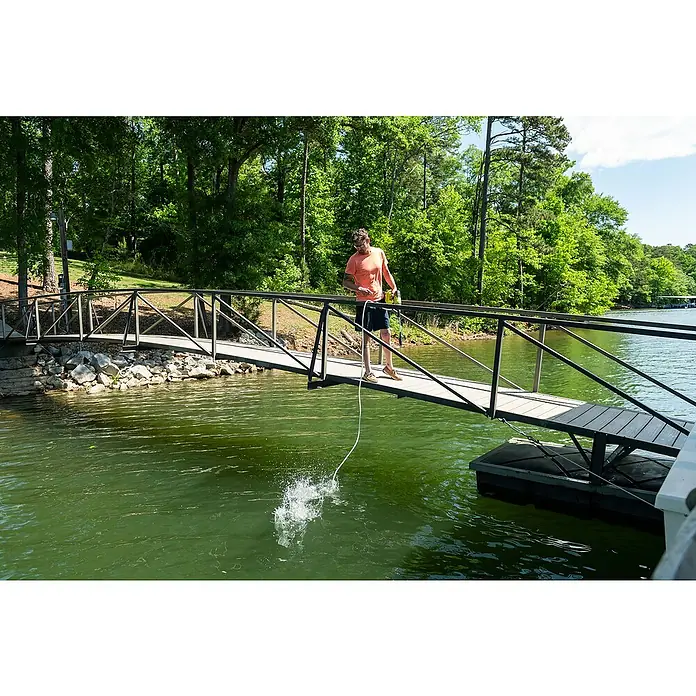 Ryobi ONE+ Akku-Hochdruckreiniger RY18PWX41A-0 - 18 V, Ohne Akku, Max. Druck: 41 bar - Ein Mann steht auf einer Brücke und wirft eine Leine ins grüne Wasser, umgeben von Bäumen und blauem Himmel.