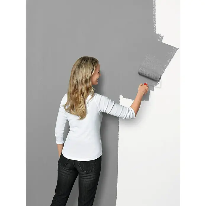 SCHÖNER WOHNEN-Farbe Trendstruktur Dekoputz-Set Sichtbeton-Optik - 3 -tlg. - Frau streicht eine Wand mit einer grauen Farbrolle, die das darunterliegende Weiß bedeckt und einen modernen Kontrast schafft.