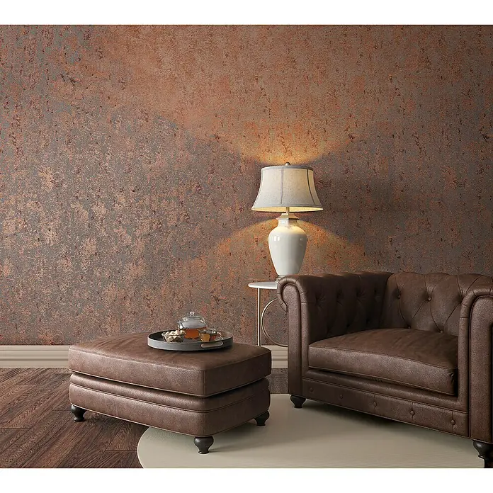 Marburg Avalon Vliestapete - Kupfer, Betonoptik, 10,05 x 0,53 m - Elegantes Wohnzimmer-Arrangement mit einem braunen Ledersessel und passendem Hocker, beleuchtet durch eine stilvolle Tischlampe.