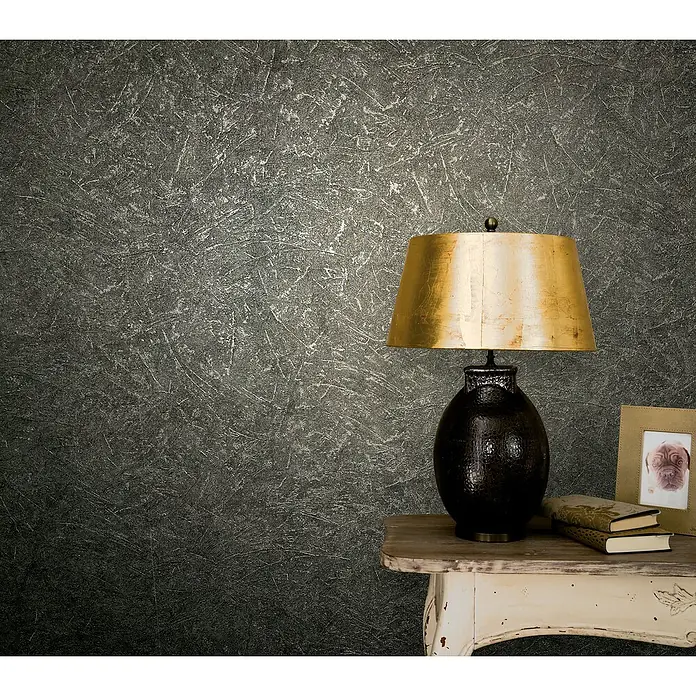 Marburg Vintage Deluxe Vliestapete - Anthrazit-Silber, Uni, 10,05 x 0,53 m - Eine Nachttischlampe mit einem goldenen Schirm steht auf einem Tisch, daneben ein Bilderrahmen und Bücher.