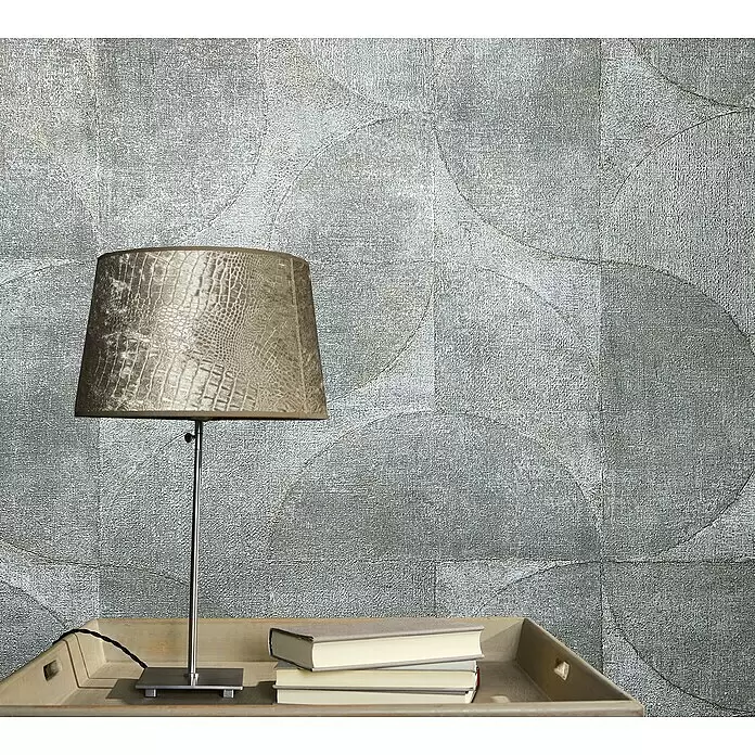Marburg Vintage Deluxe Vliestapete - Blau-Grau, Grafisch, 10,05 x 0,53 m - Eine metallene Tischlampe mit strukturiertem beigem Lampenschirm steht auf einem Holztablett mit Büchern vor einer silbernen Mustertapete.