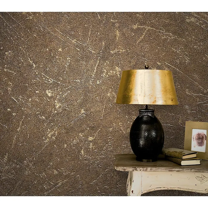 Marburg Vintage Deluxe Vliestapete - Gold, Uni, 10,05 x 0,53 m - Eine dekorative Tischlampe mit goldenem Schirm und Büchern auf einem kleinen Tisch, vor einer strukturierten Wand.