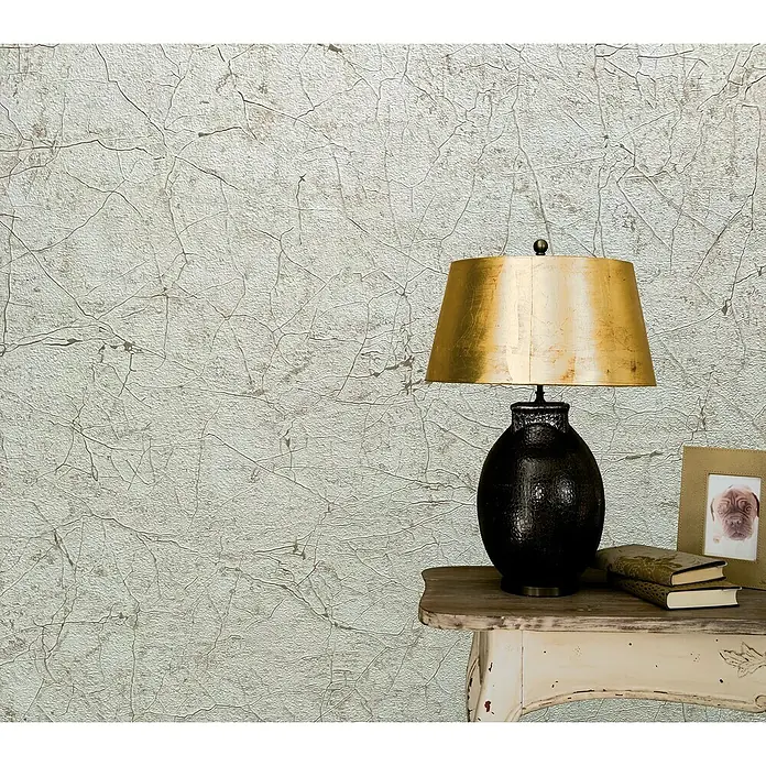 Marburg Vintage Deluxe Vliestapete Craquelée-Risse - Beige, Grafisch, 10,05 x 0,53 m - Elegante Tischlampe mit goldenem Schirm und dunklem Fuß auf einem Beistelltisch, ergänzt durch Bücher und einen Bilderrahmen.
