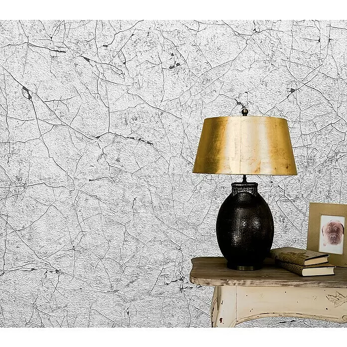 Marburg Vintage Deluxe Vliestapete Craquelée-Risse - Lichtgrau, Grafisch, 10,05 x 0,53 m - Eine goldfarbene Tischlampe mit dunklem, strukturiertem Fuß auf einem verwitterten Holztisch, neben Büchern und einem Bilderrahmen, vor einer weißen rissigen Wand.