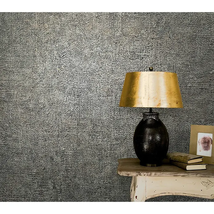 Marburg Vintage Deluxe Vliestapete - Grau-Anthrazit, Uni, 10,05 x 0,53 m - Stimmungsvolle Aufnahme einer goldenen Lampenkollektion auf einem Beistelltisch mit Büchern und einem Bilderrahmen.