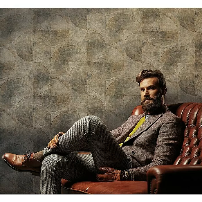 Marburg Vintage Deluxe Vliestapete - Kupfer-Anthrazit, Grafisch, 10,05 x 0,53 m - Mann in Tweed-Jackett, gelbem Pullover, gemusterten Hosen, braunen Schuhen und Lederhandschuhen sitzt auf einem Ledersessel.