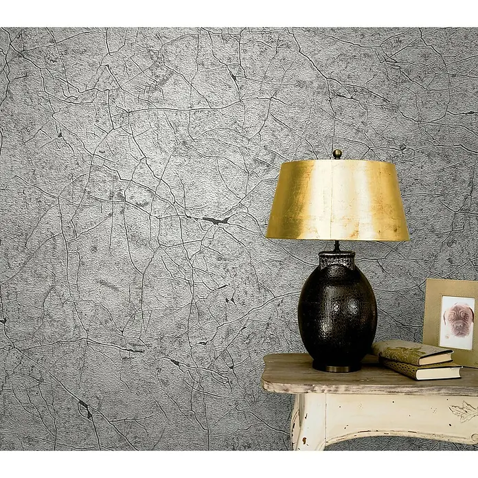Marburg Vintage Deluxe Vliestapete Craquelée-Risse - Grau, Grafisch, 10,05 x 0,53 m - Elegante Nahaufnahme einer Tischlampe mit goldenem Schirm, stilvollen Büchern und einem Fotorahmen auf einem dekorierten Beistelltisch.