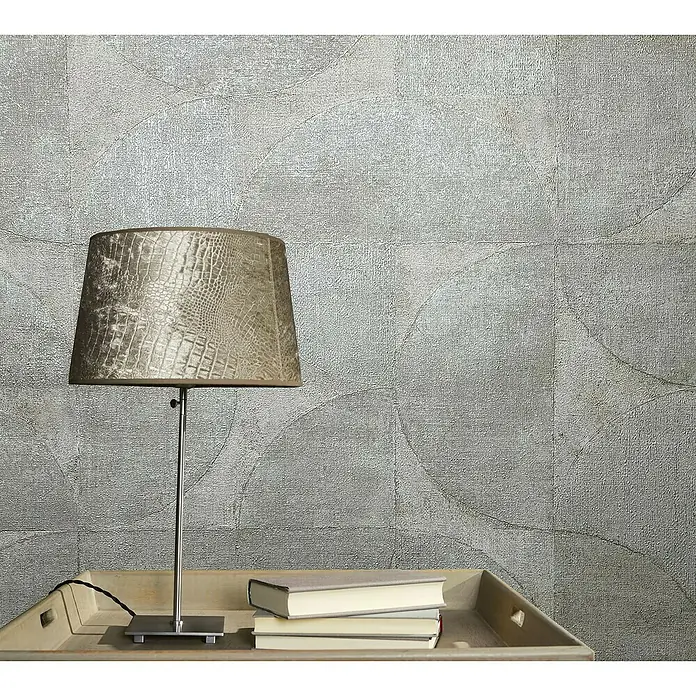Marburg Vintage Deluxe Vliestapete - Beige-Pearl, Grafisch, 10,05 x 0,53 m - Eine moderne Lampe mit Schirm in Kroko-Optik steht auf einem Tisch mit Büchern. Das Zimmer hat eine strukturierte Tapete.