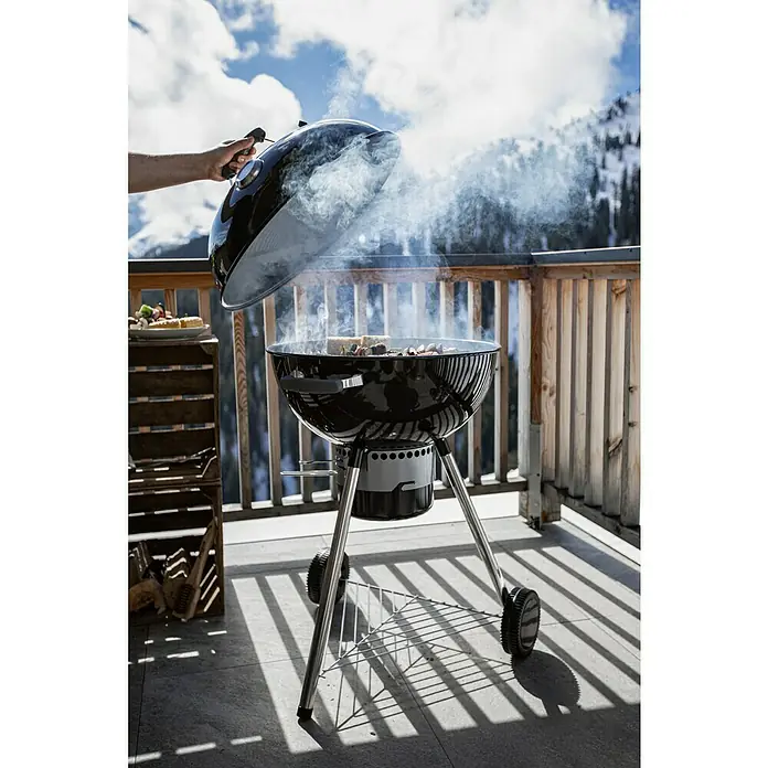 Kingstone Kugelgrill Bullet 57 - Durchmesser Grillfläche: 57 cm, Schwarz - Ein schwarzer Kugelgrill mit geöffnetem Deckel, aus dem Rauch aufsteigt, auf einer Terrasse mit Blick auf die Berge.