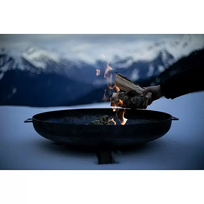 Feuerschale - Durchmesser: 80 cm, Eisen - Hand legt Holz in eine dunkle Metall-Feuerschale mit Flammen in verschneiter Berglandschaft.