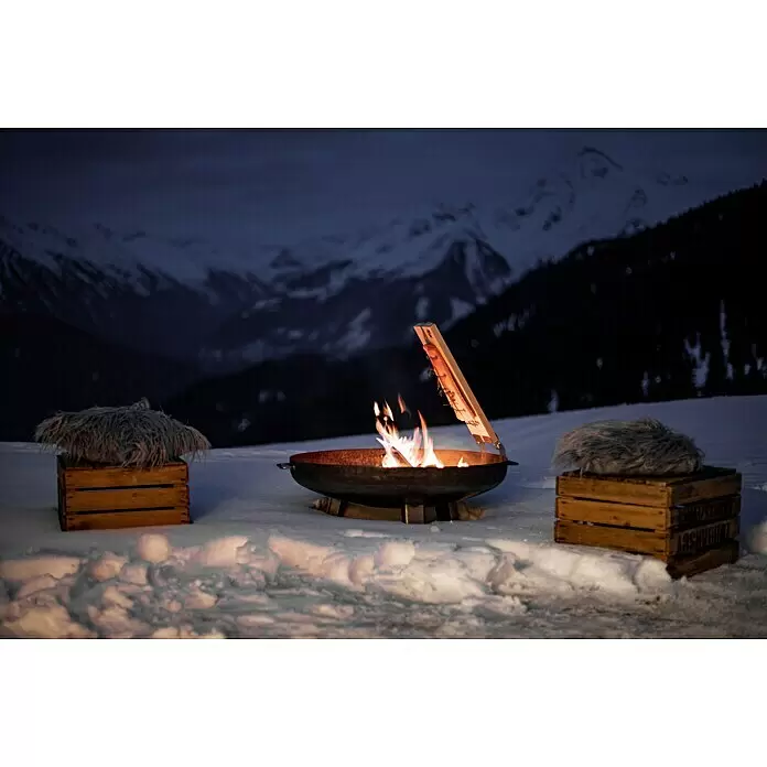 Feuerschale - Durchmesser: 80 cm, Eisen - Dunkle Metallfeuerschale mit brennendem Feuer, zum Garen von Lachs auf einem Holzbrett, in verschneiter Berglandschaft bei Nacht.