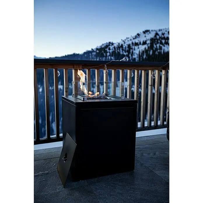 MWH Feuerstelle Palermo - 55 x 55 x 79 cm, Aluminium, Schwarz - Ein moderner, schwarzer Gas-Feuertisch mit Glasaufsatz steht auf einer Terrasse, im Hintergrund eine verschneite Berglandschaft bei Dämmerung.