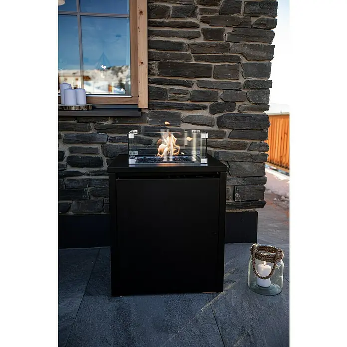 MWH Feuerstelle Palermo - 55 x 55 x 79 cm, Aluminium, Schwarz - Nahaufnahme eines Bioethanol-Feuertisches im Außenbereich mit einem dekorativen Glasgefäß und einer Steinmauer im Hintergrund.